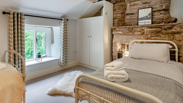 A bedroom with a stone wall, a bed with a striped pillow, a white wardrobe, and a window with curtains. - 2 Bed in Hope Valley (oc-88979) (Sheffield)