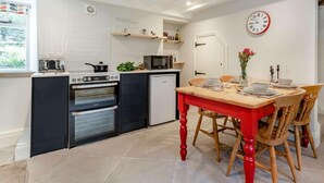 A compact kitchen with a stove, oven, microwave, and a small refrigerator. There is a dining table with chairs and a clock on the wall. - 2 Bed in Hope Valley (oc-88979) (Sheffield)