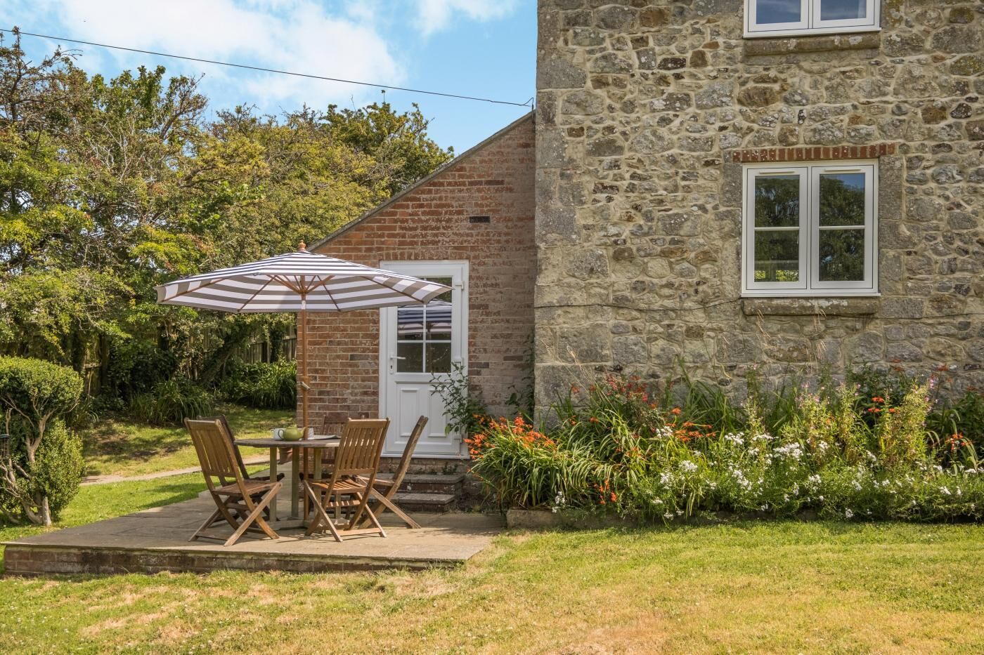 A stone building with a brick roof, a patio with a table and chairs, and a garden with various plants and flowers.