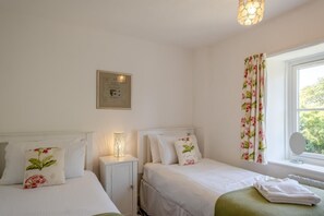 A bedroom with two beds, a bedside table with a lamp, a framed picture on the wall, and a window with curtains. - 3 Bed in Ventnor (oc-ic179) (Godshill)