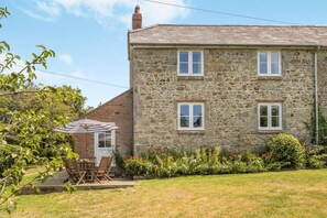 A stone building with a brick extension, a patio with a table and chairs, and a garden with various plants. - 3 Bed in Ventnor (oc-ic179) (Godshill)