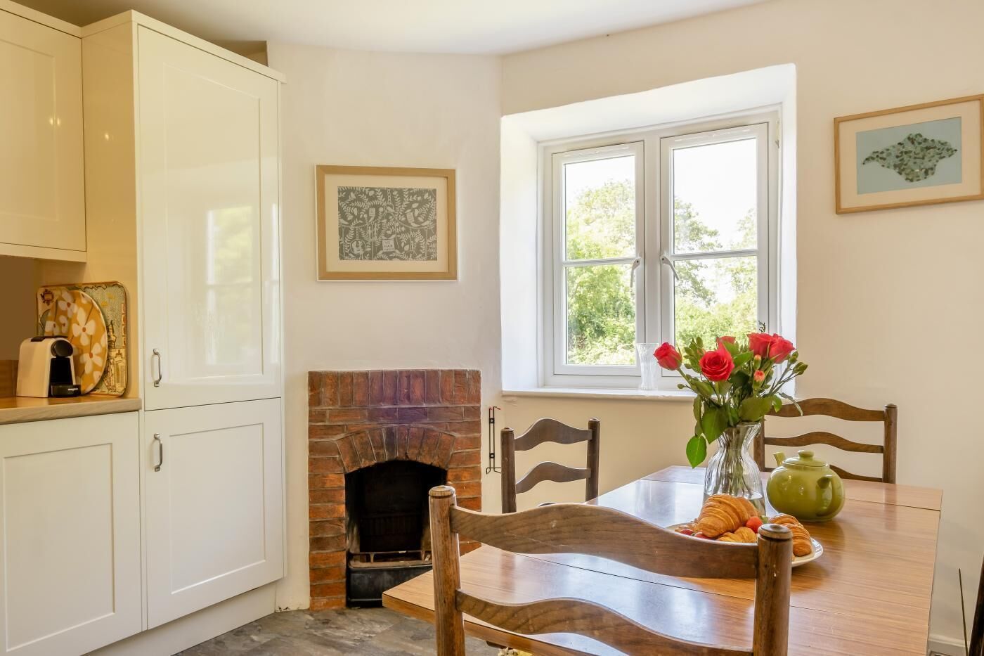 A dining area with a brick fireplace, white cabinetry, a table with chairs, and a window with a view of greenery.