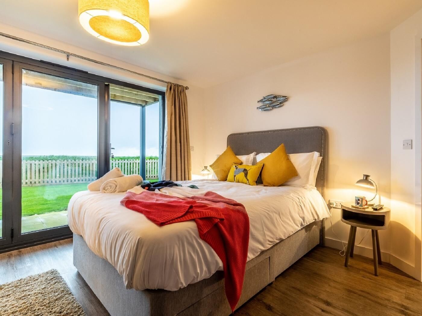 Een slaapkamer met een bed, nachtkastje, lamp, raam met gordijnen en uitzicht op een hek.