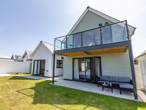 A modern two-story house with a balcony, a sliding glass door entrance, and a patio area with outdoor furniture. - 4 Bed in Bideford (oc-75706) (Northam)