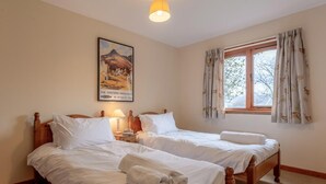 Ein Hotelzimmer mit zwei Holzbetten, einem Nachttisch mit Lampe, einem Fenster mit Vorhängen und einem Gemälde der schottischen Highlands.
