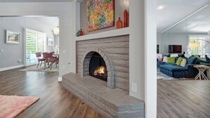 Een moderne woonkamer met een open haard, een eethoek met een tafel en stoelen, en een groot raam met zonwering.