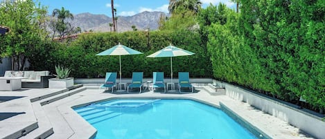 A swimming pool with lounge chairs and umbrellas, surrounded by greenery and mountains in the background.
