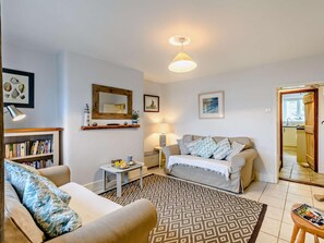 Ein Wohnzimmer mit einer Couch, einem Couchtisch, einem gemusterten Teppich, einem Bücherregal, einem Spiegel und einer zur Küche hin führenden Tür.