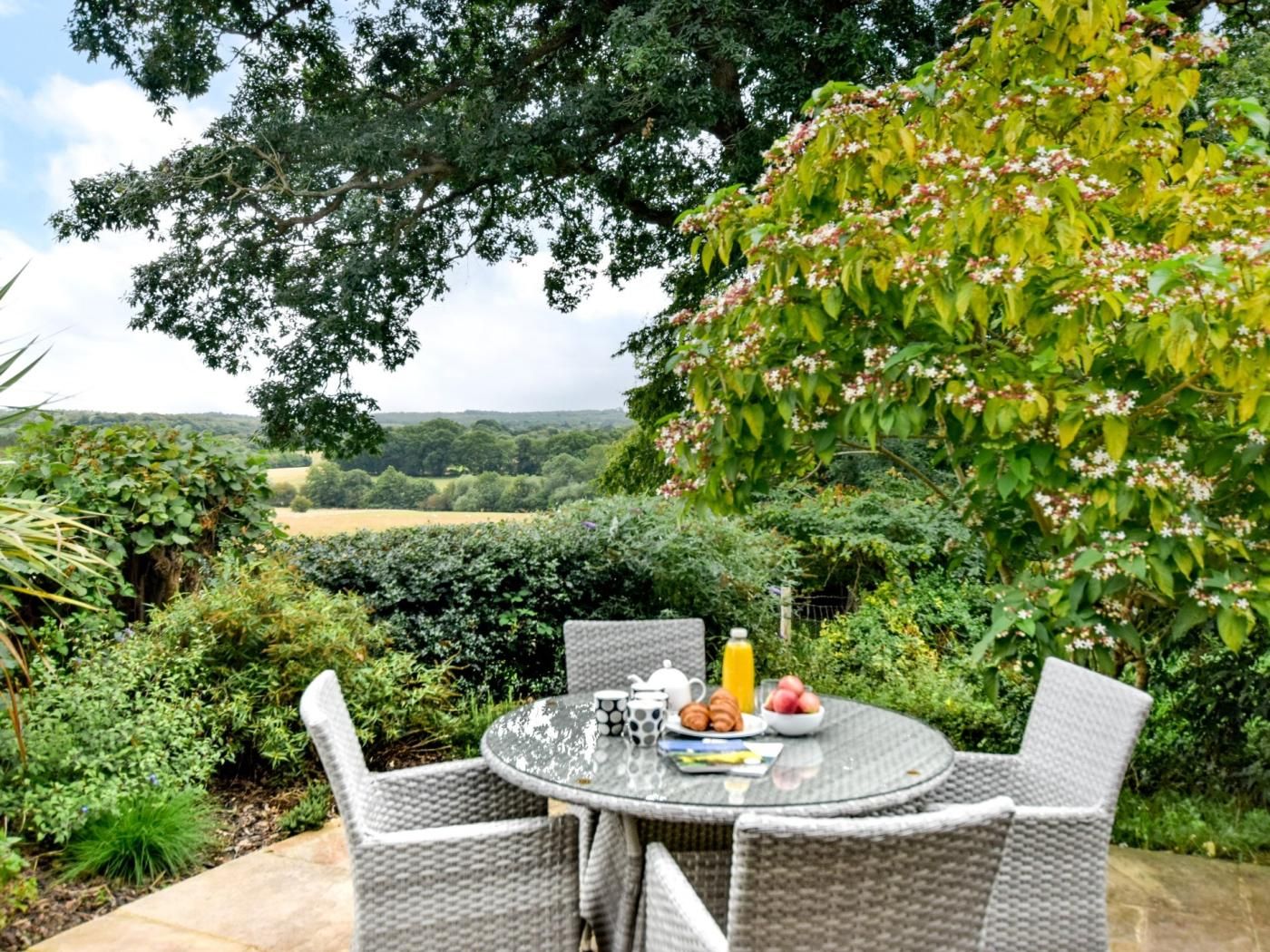 Une table ronde d’extérieur entourée de chaises en osier, une théière, une tasse, des croissants et des fruits.