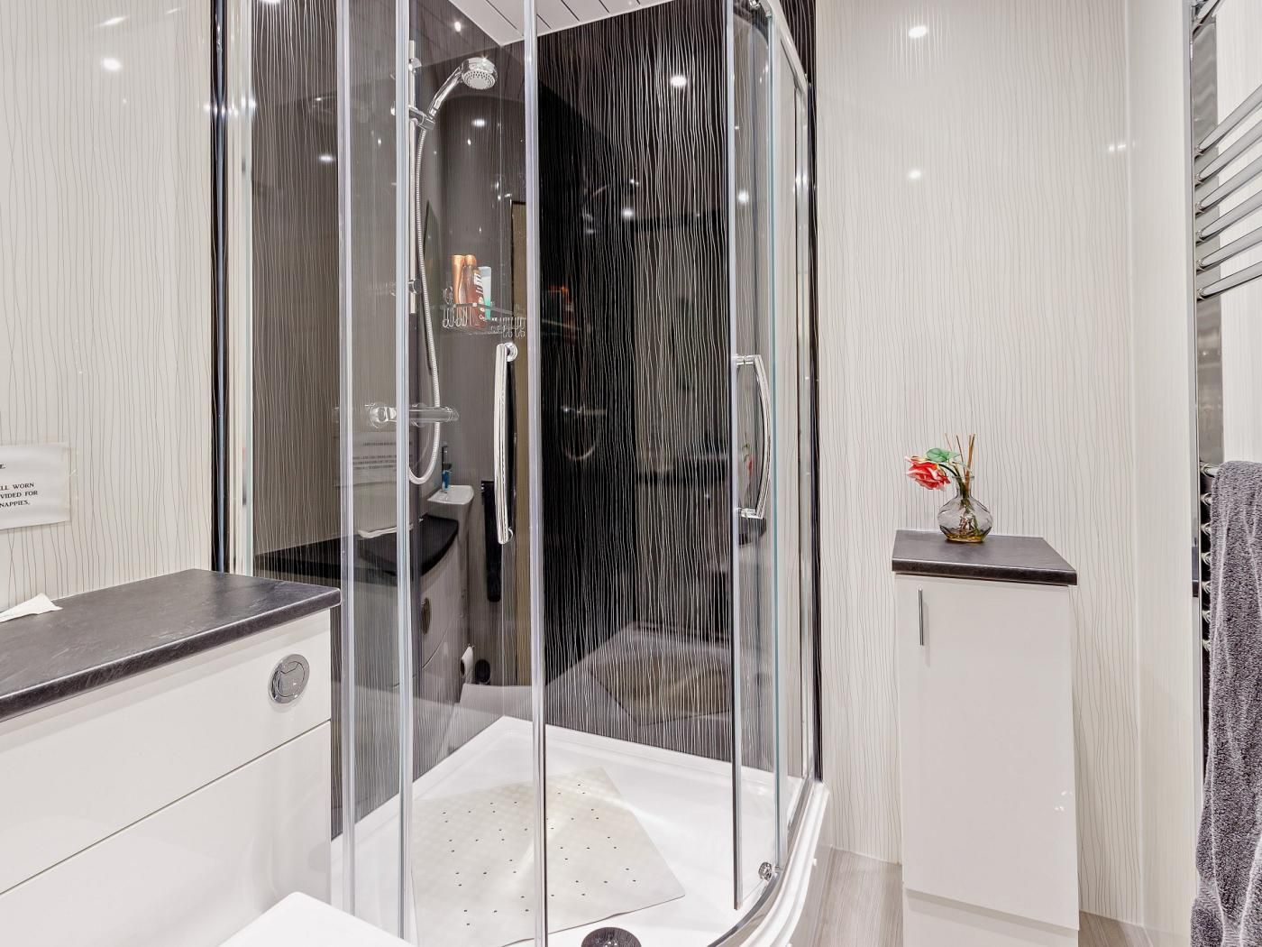 A modern bathroom with a glass-enclosed shower, a vanity with a dark countertop, and a small shelf with a vase.