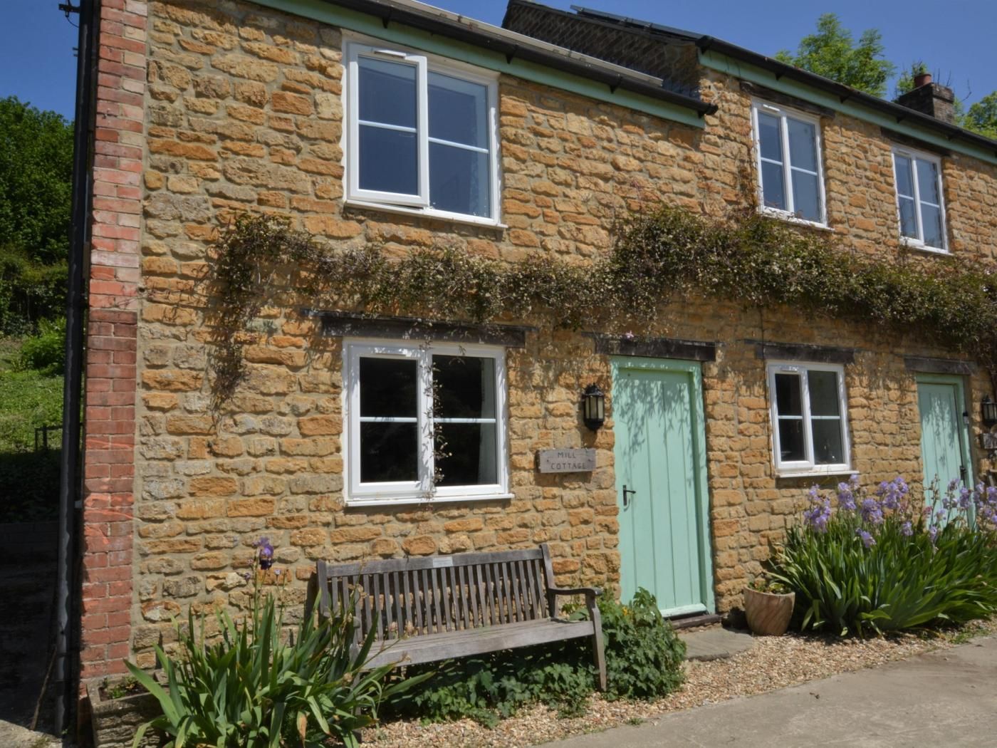 A stone cottage with a bench, a green door, and a sign that reads 'Mill Cottage.'