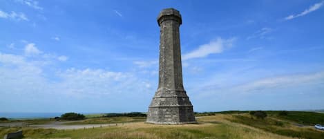 Ein hohes, schmales Denkmal mit flacher Spitze, umgeben von grünen Feldern und blauem Himmel.