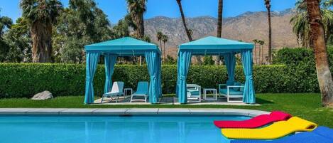 A pool area with two gazebos, lounge chairs, and a clear view of mountains in the background.
