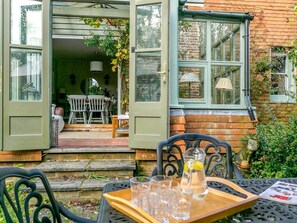 Eine offene Tür führt in einen Essbereich mit Tisch und Stühlen, ein Fenster mit Lampe und eine Ziegelwand.