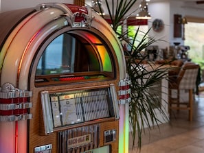 A jukebox with a wooden finish and chrome accents, located in a room with a dining area and a plant.