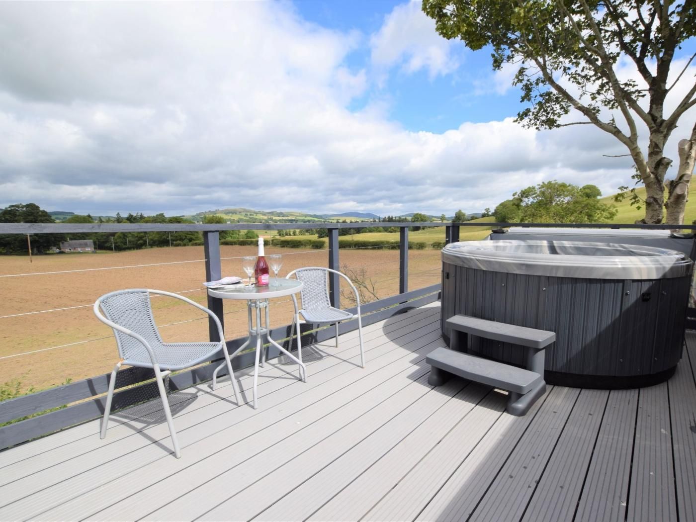 A hot tub on a wooden deck with two chairs and a table, overlooking a field.