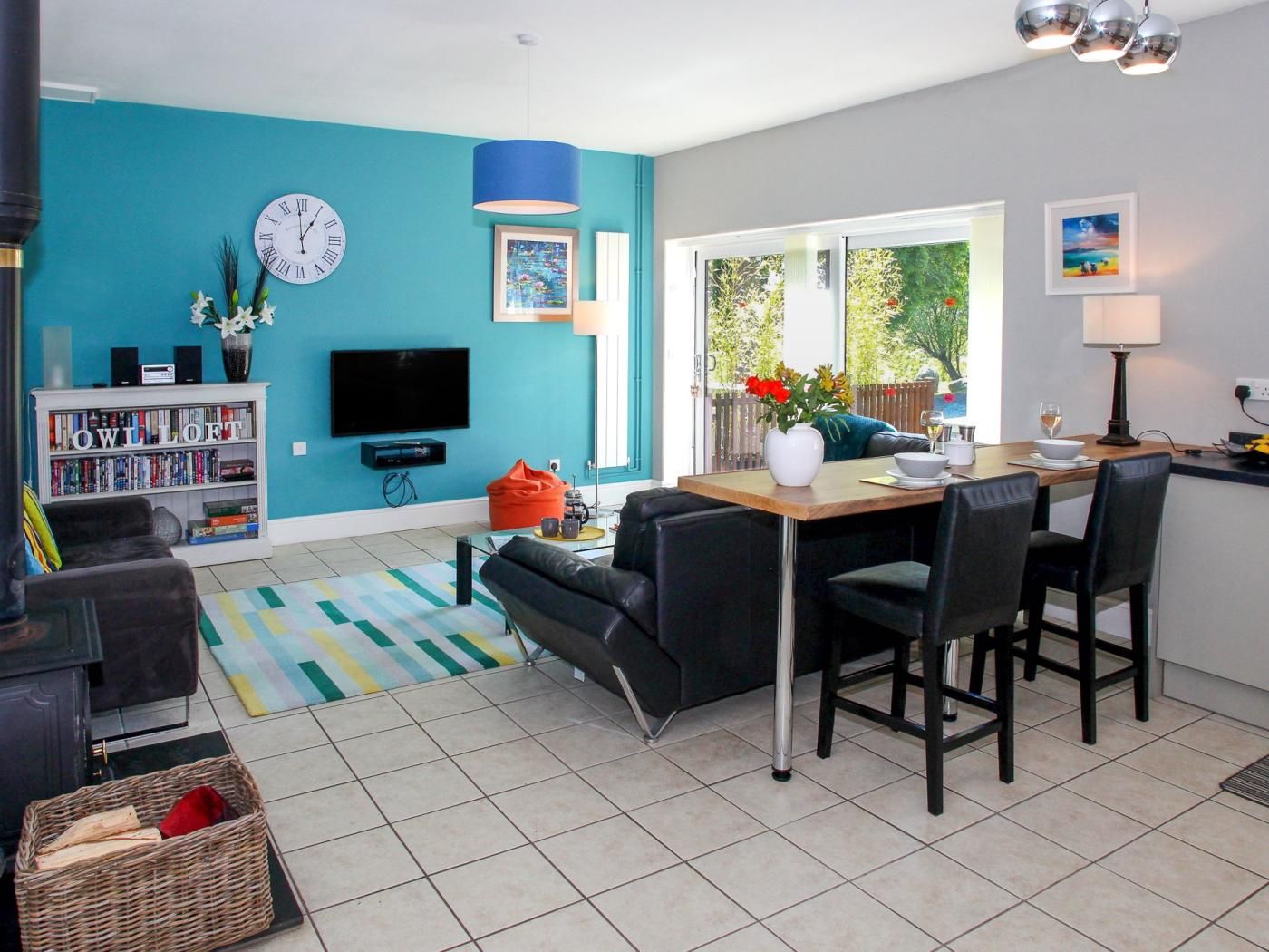 A modern living room with a teal accent wall, a flat-screen TV, a bookshelf, a dining area with a table and chairs, and a large window with a view of greenery.