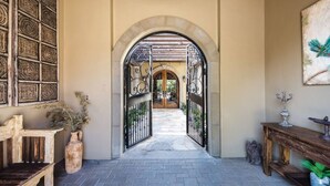 An arched entrance with a metal gate, a wooden bench, a vase with dried plants, and a small table with a candelabra. - 5 BR Custom Mediterranean Estate In Andreas Hills, 5 King Beds Putting Green... (Palm Springs)