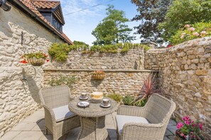A stone wall with a small table and two wicker chairs, a hanging flower basket, and a stone wall with climbing plants.