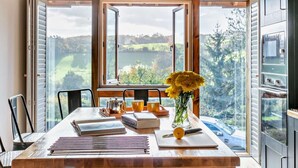 Un comedor con una mesa dispuesta para dos, un florero con flores amarillas y vistas a un lago y árboles a través de ventanas amplias.