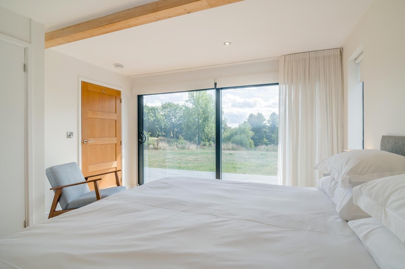 Une chambre avec un grand lit, une chaise, une porte en bois et une vue sur un espace verdoyant, visible à travers une porte-fenêtre coulissante.