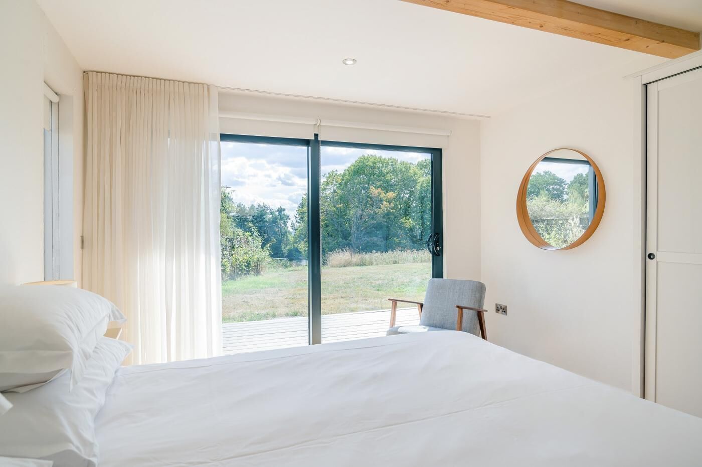 Ein Schlafzimmer mit einem Bett, einem Sessel, einem runden Spiegel und einer Glastür zum Außenbereich.