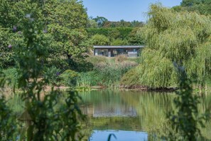 A lakeside property with a modern building, surrounded by lush greenery and a weeping willow. - 3 Bed in Tiverton (oc-59634) (Whitnage)