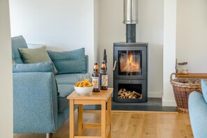 A cozy living room with a blue sofa, a small wooden table with snacks and drinks, and a wood-burning stove. - 3 Bed in Tiverton (oc-59634) (Whitnage)