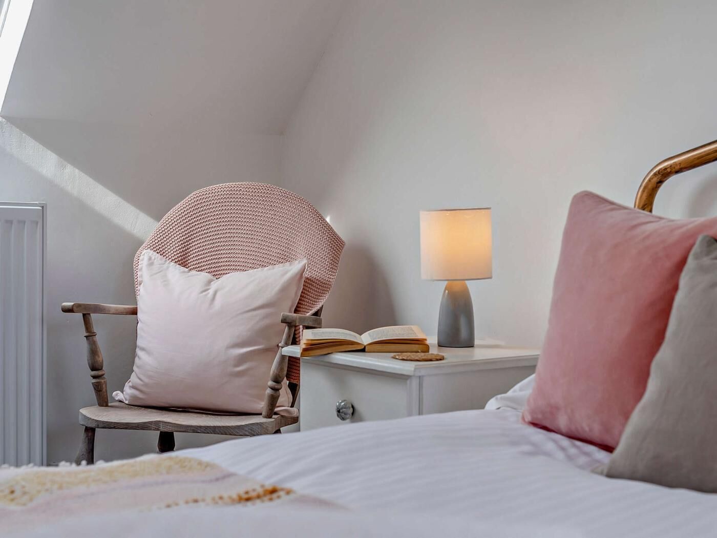 Ett sovrum med ett sluttande tak, en trästol med en kudde, ett sängbord med en lampa och en säng med kuddar.