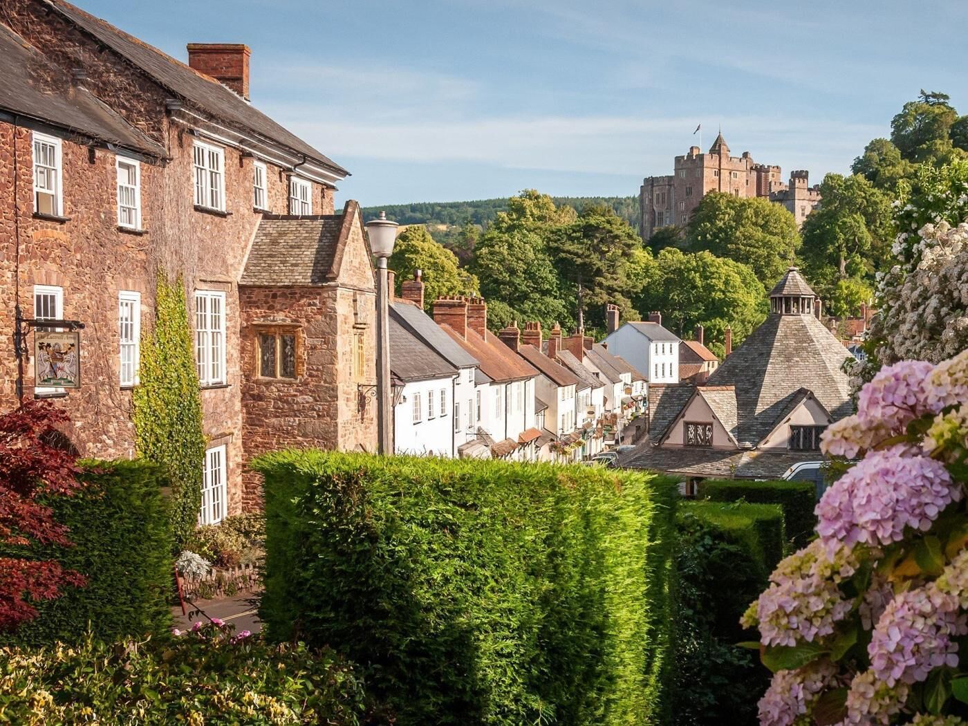 Ein historisches Steinhaus mit traditioneller Architektur, ein weißes Gebäude mit Satteldach und eine Burg im Hintergrund.