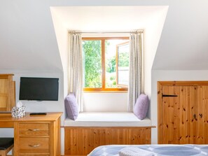 A bedroom with a sloped ceiling, a window seat with cushions, a bed, a wooden wardrobe, and a television on a wooden dresser. - 1 Bed in Taunton (oc-ndown) (Otterford)