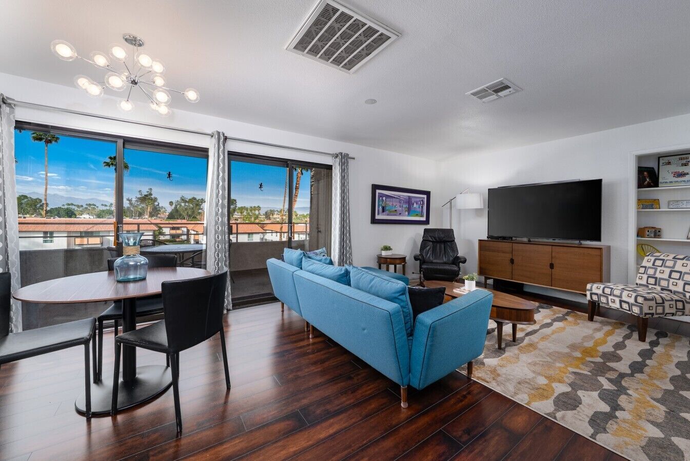 A modern living room with a blue sofa, a dining table, a TV, and a large window offering a view of palm trees and buildings.