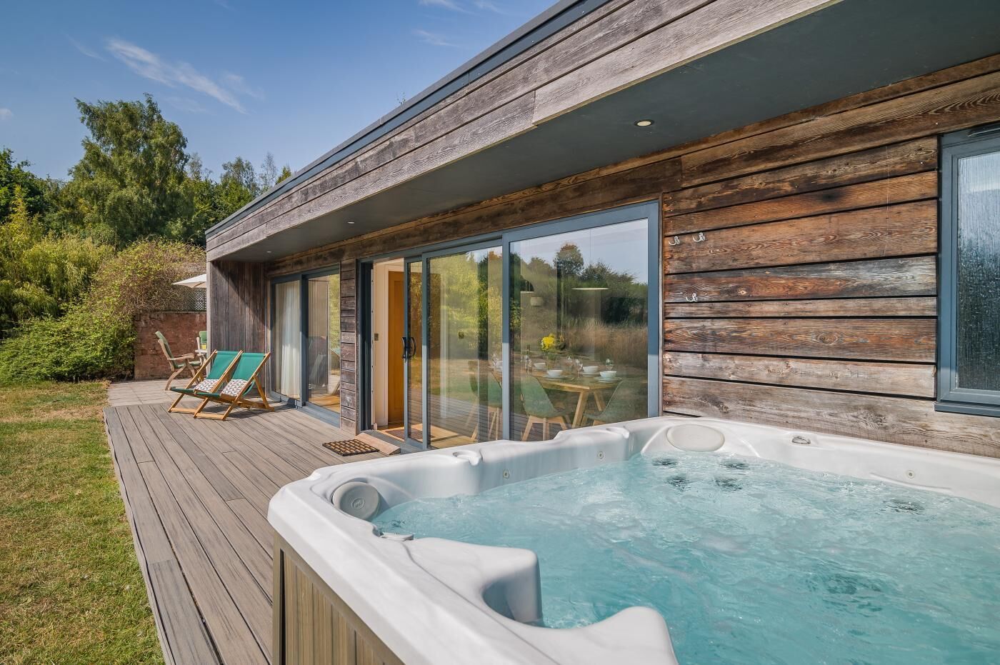 A modern outdoor hot tub area with wooden decking, lounge chairs, and a glass-enclosed patio.