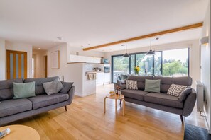 A modern living room with two sofas, a coffee table, and a kitchen in the background. - 3 Bed in Tiverton (oc-59635) (Whitnage)
