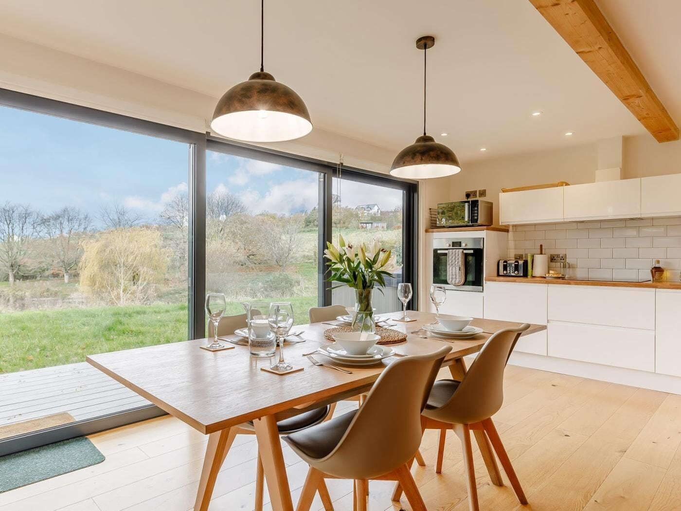 Une cuisine moderne dotée de grandes fenêtres, un coin repas avec une table et des chaises en bois, et une vue sur l’extérieur.