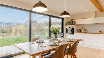 Une cuisine moderne dotée de grandes fenêtres, un coin repas avec une table et des chaises en bois, et une vue sur l’extérieur.