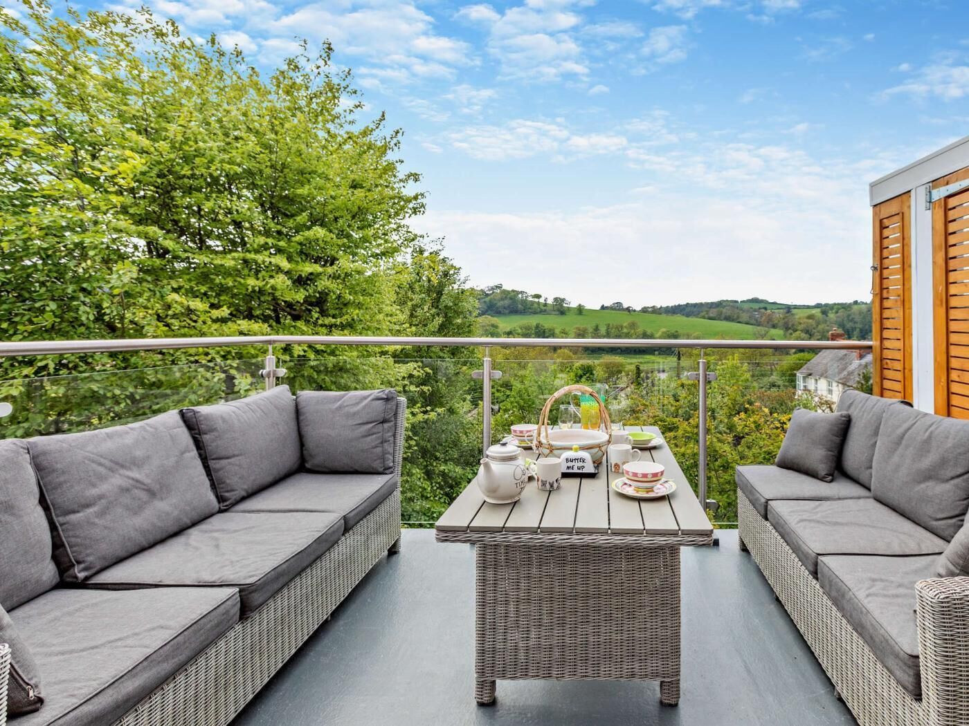 A balcony with wicker furniture, a table set for tea, and a scenic view of green hills.