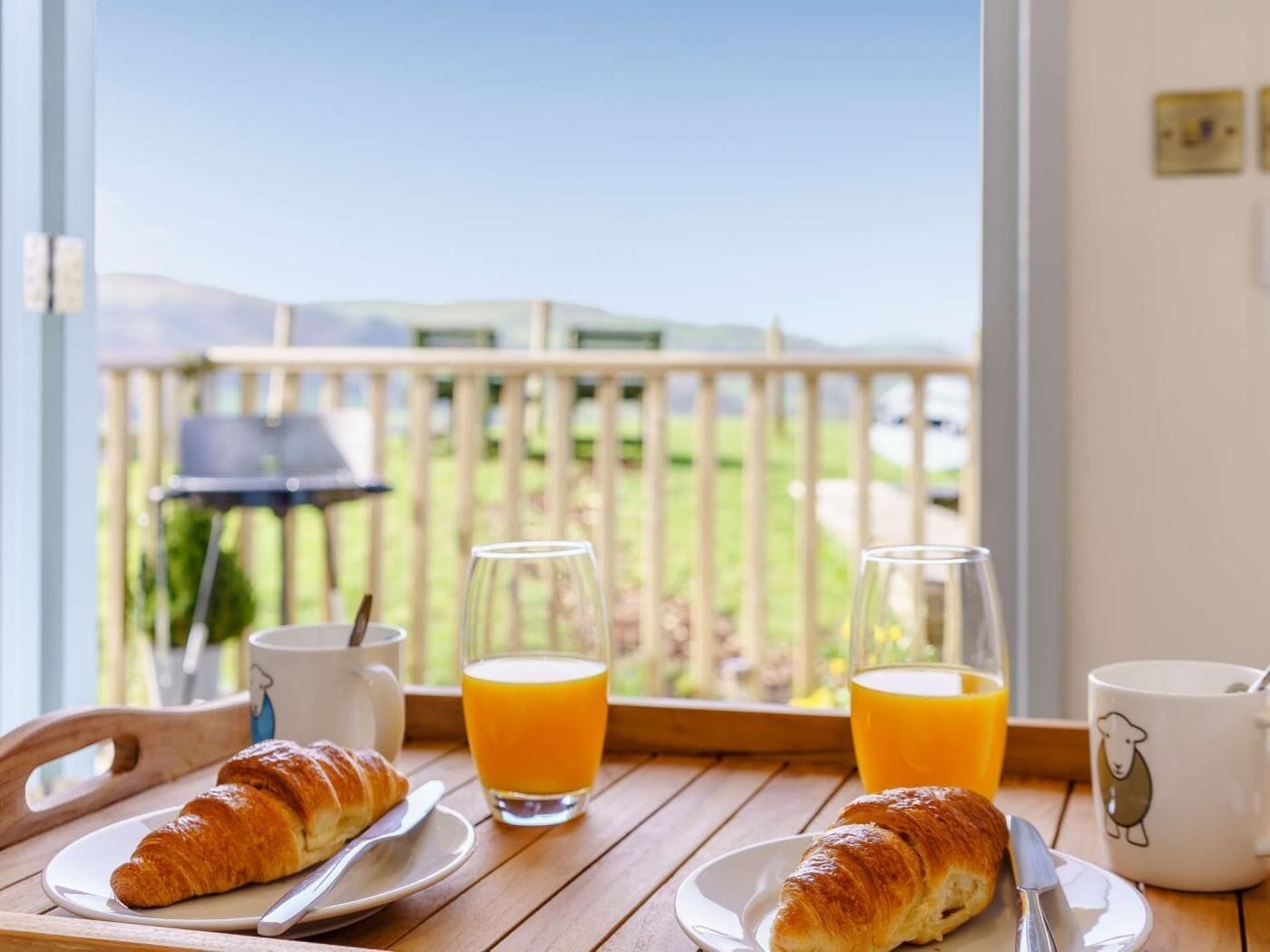 A breakfast table with croissants, juice glasses, and a mug, overlooking a balcony with a view of a green landscape.