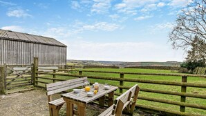 Ein rustikaler Sitzbereich im Freien mit Tisch und Bänken, einem Holzzaun und Blick auf eine ländliche Landschaft.