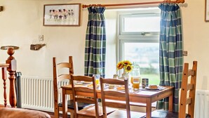 Ein Essbereich mit einem Tisch für zwei Personen, Holzstühlen, einer Heizung, einem Fenster mit Karottenvorhängen und einem gerahmten Bild an der Wand.