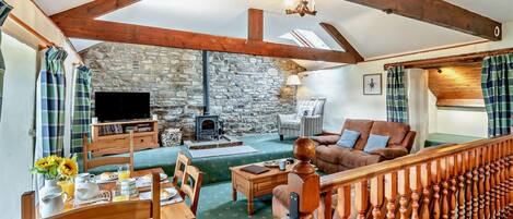 A cozy living room with a stone wall, wooden beams, a fireplace, a sofa, a TV, and a dining area with a table set for two.