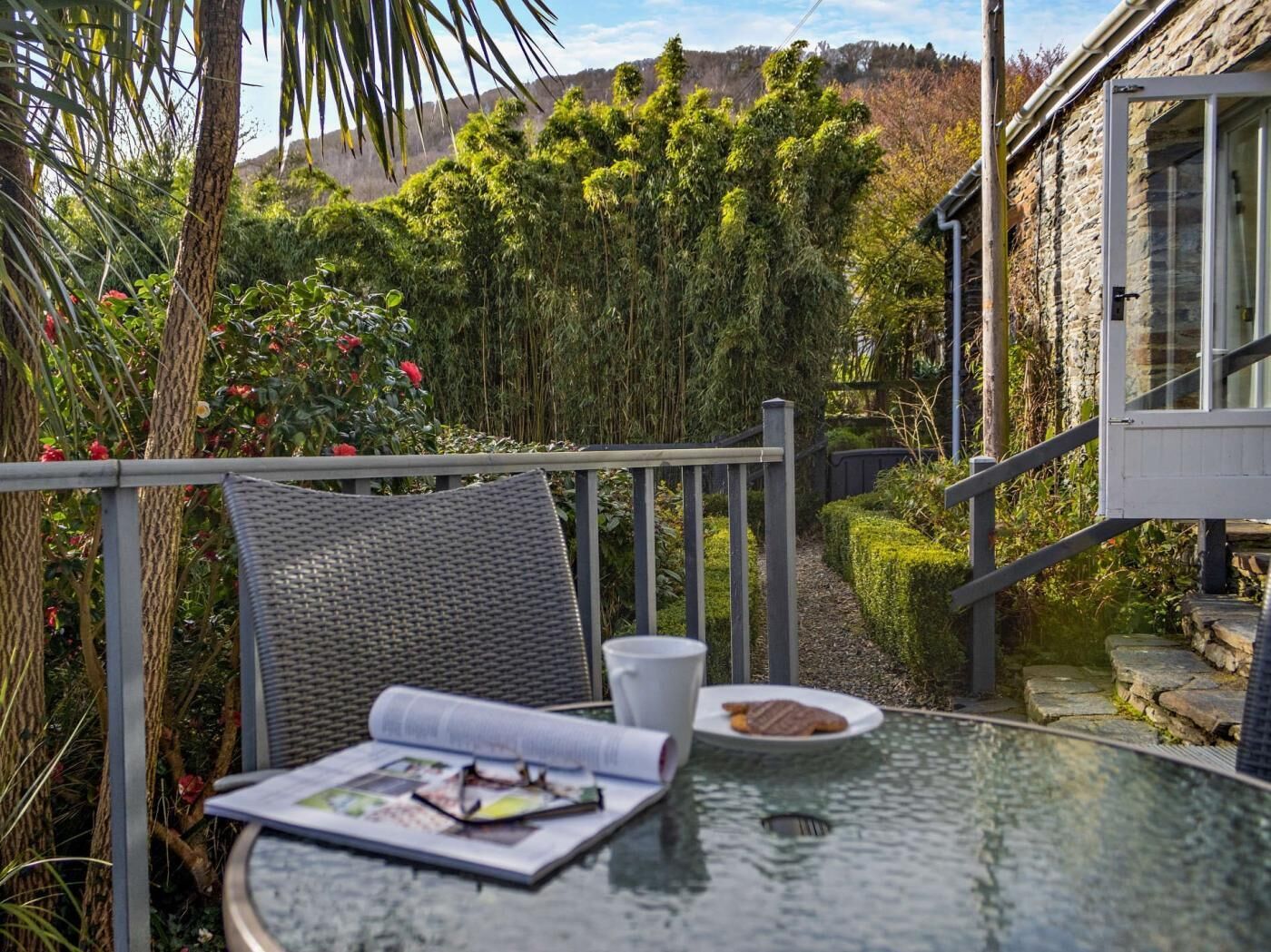 A glass table with a magazine, a cup, and a plate with food, set against a backdrop of greenery and a stone building.