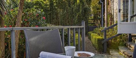 A glass table with a magazine, a cup, and a plate with food, set against a backdrop of greenery and a stone building.