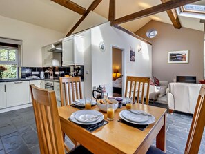 A dining area with a wooden table set for two, a kitchen in the background, and a living area with a sofa and TV. - 2 Bed in Aberdyfi (oc-dy031) (Aberdovey)
