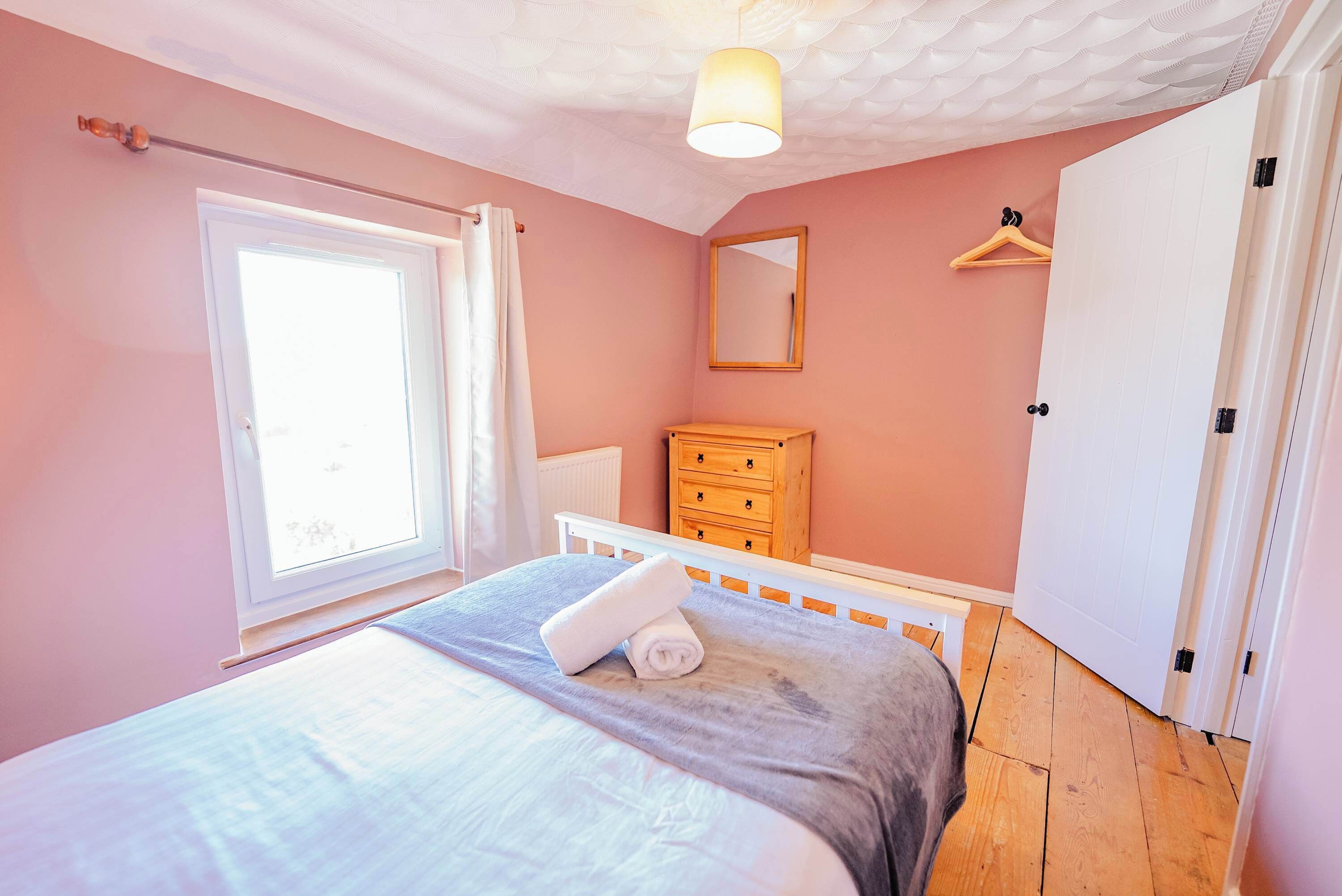 Ein Schlafzimmer mit Bett, Kommode, Spiegel, Kleiderstange und Fenster.