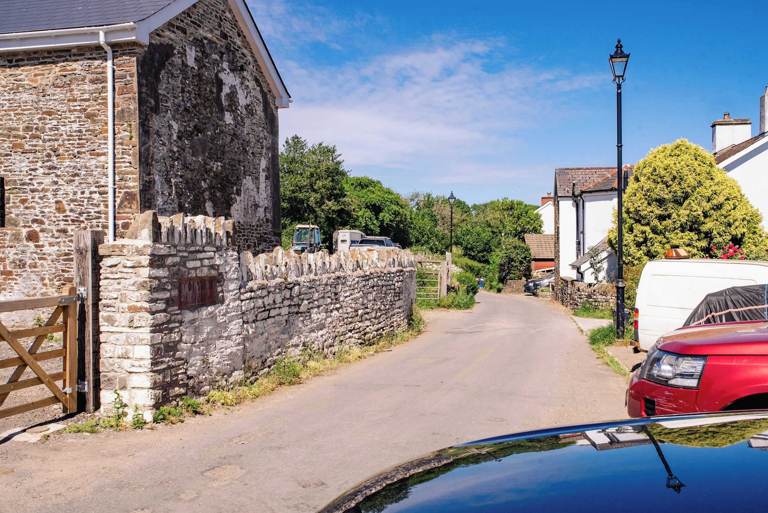 A narrow rural road with stone walls and a wooden gate, leading to a stone building with a white door.