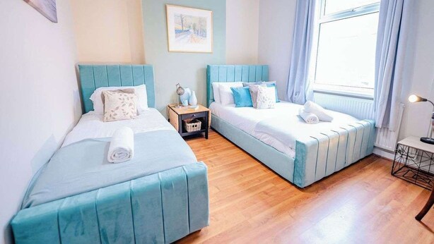 Una habitación con dos camas, una mesita, una mesilla de noche, una ventana con cortinas y un cuadro en la pared.