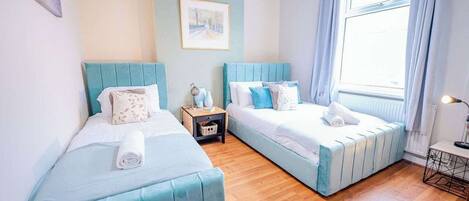 A room with two beds, a small table, a nightstand, a window with curtains, and a painting on the wall.