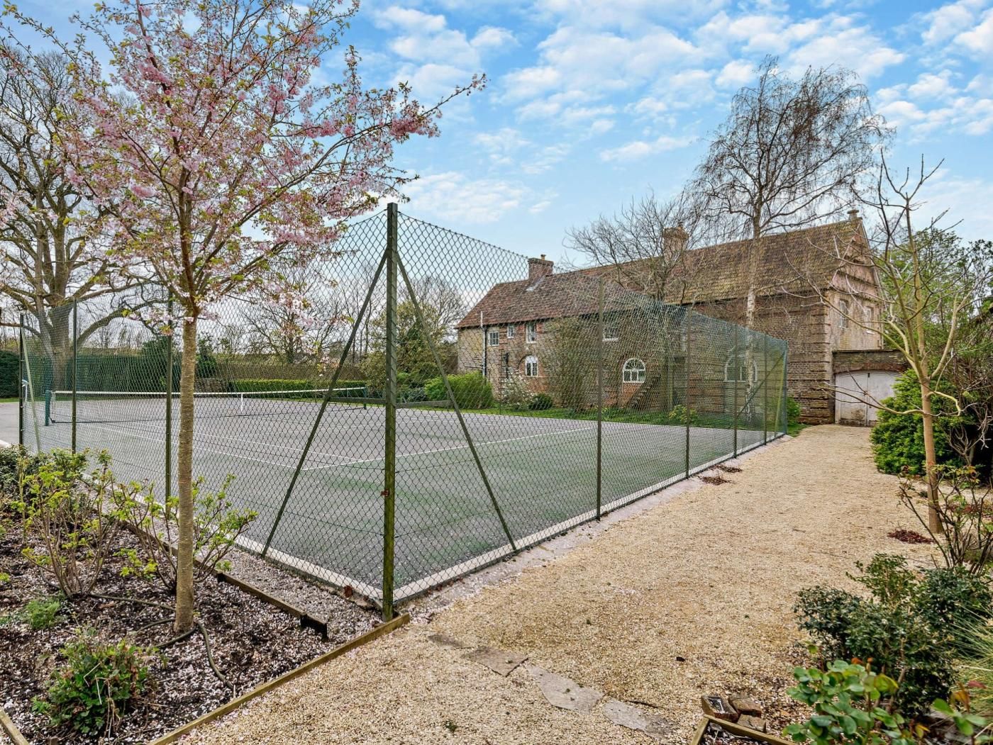 A tennis court enclosed by a chain-link fence, surrounded by a gravel path and a brick building.