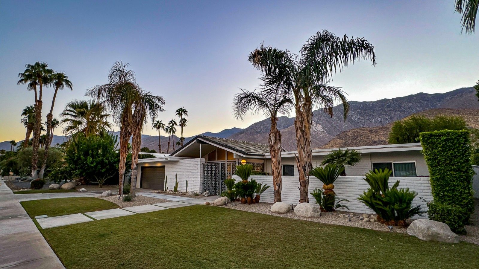 A modern house with a garage, surrounded by palm trees and a mountain backdrop.